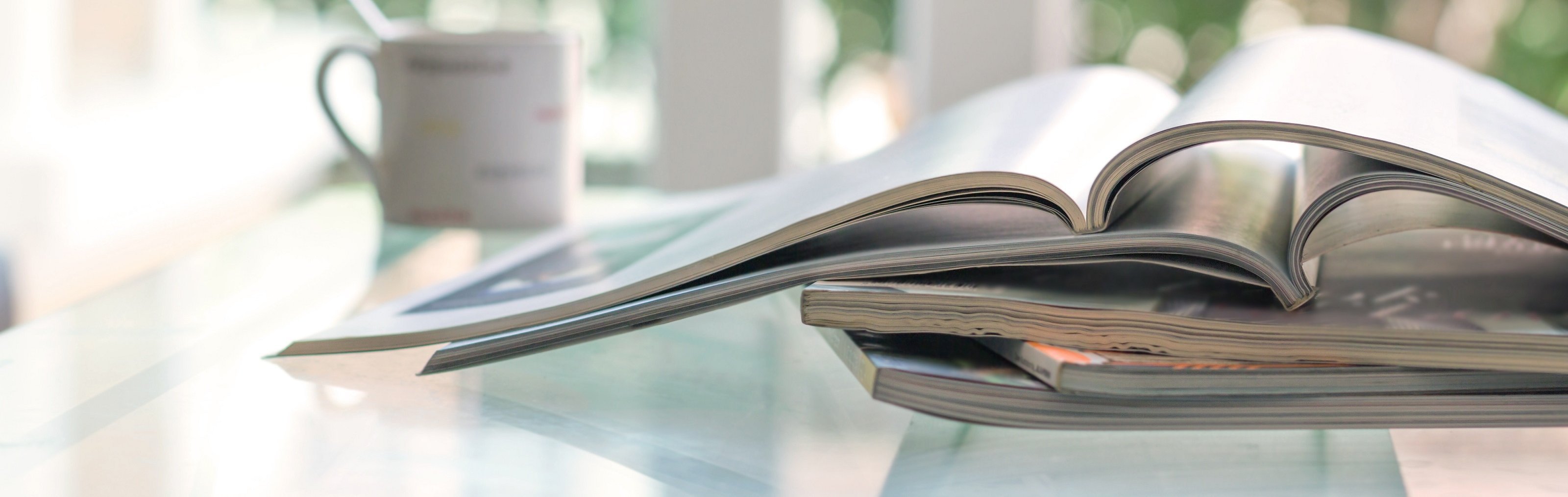 Das Foto zeigt einen Stapel an Magazinen und Zeitungen, das oberste ist aufgeschlagen. Im Hintergrund links ist eine Kaffeetasse erkennbar. 