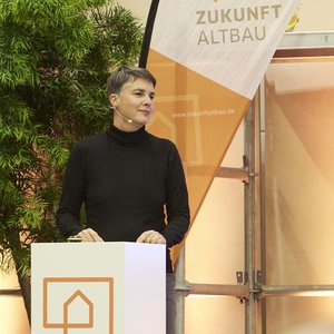 Referentin Sabine Erber bei ihrem Vortrag auf der Bühne. Im Vordergrund ist zudem ein Kameramann von hinten zu sehen, der die Szene für den Livestream mitfilmt. 