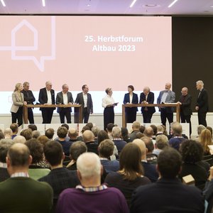 Diskussionsrunde auf der Bühne an zwei großen Tischen mit elf Personen. 