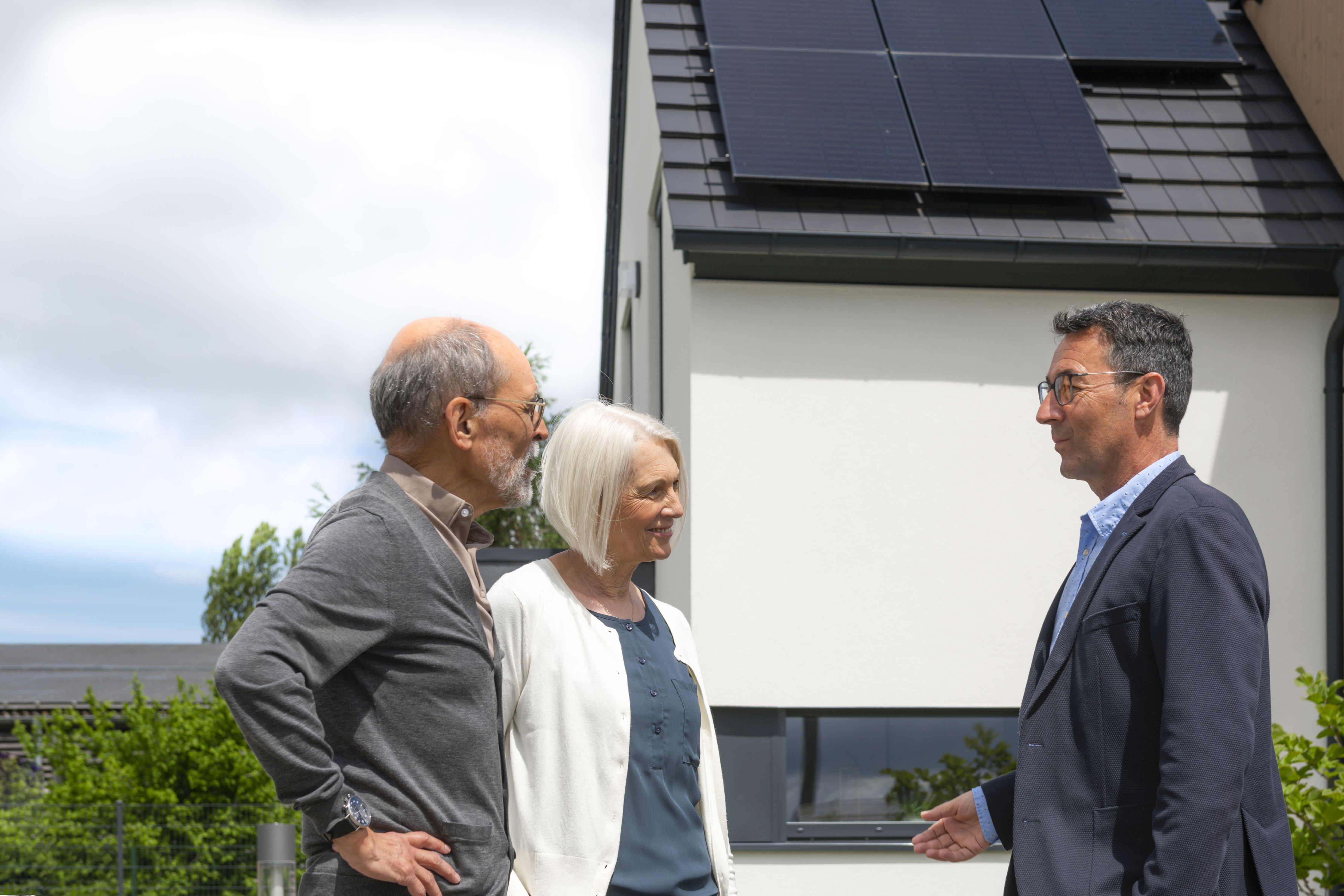 Auf dem Bild sieht man zwei Männer und eine Frau. Ein Mann steht auf der rechten Seite und spricht mit dem anderen Mann und der Frau. Sie stehen ihm gegenüber. Im Hintergrund sind Teile von Solarmodulen zu sehen