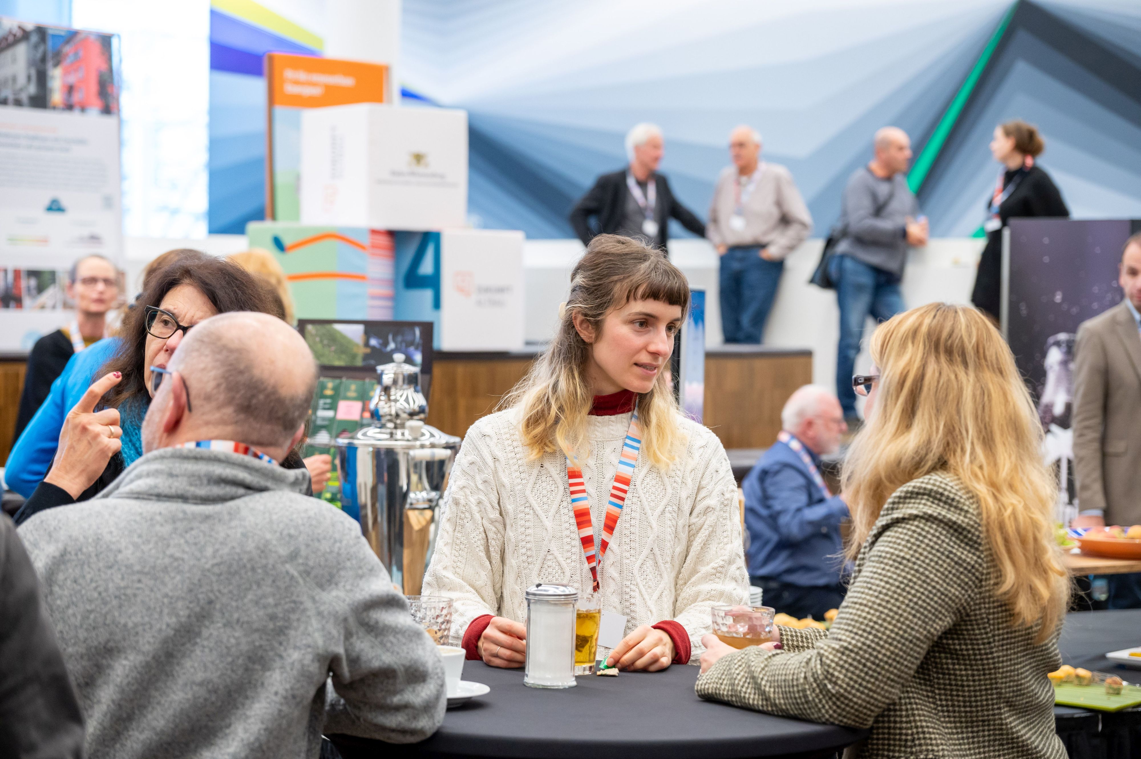 Vier Personen stehen rund um einen Stehtisch und tauschen sich angeregt aus. Im Hintergrund sieht man Personen auf einer Erhöhung, auch sie unterhalten sich miteinander.