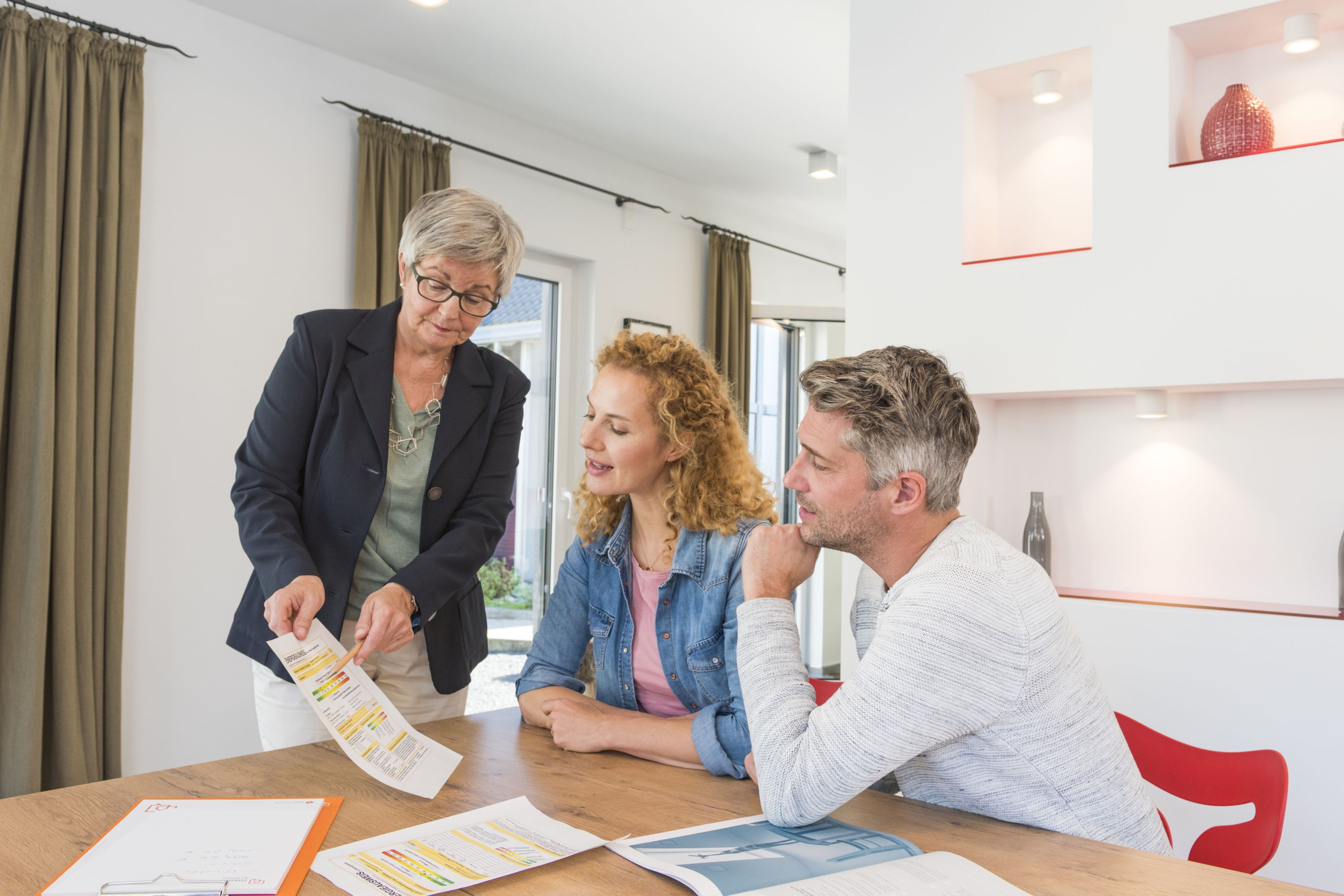 Eine Energieberaterin links im Bild zeigt einem Paar rechts im Bild ein Informationsblatt. Sie sitzen an einem Esstisch, im Hintergrund sieht man das Wohnzimmer.
