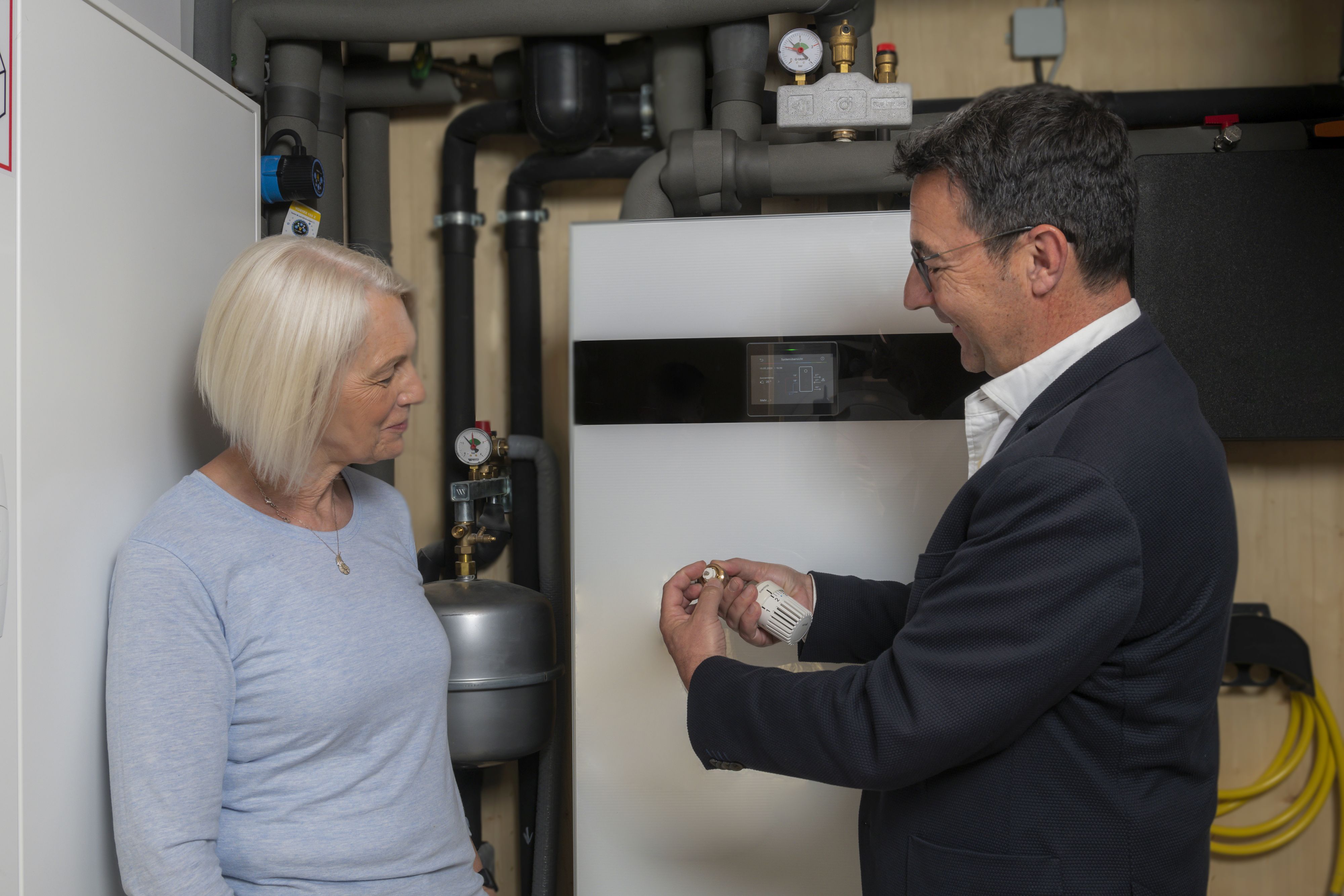 Im Bild sind eine Frau und ein Mann zu sehen. Sie stehen im Keller vor einer Heizung. Der Mann im Anzug auf der rechten Seite zeigt der Dame ein Thermostatventil.