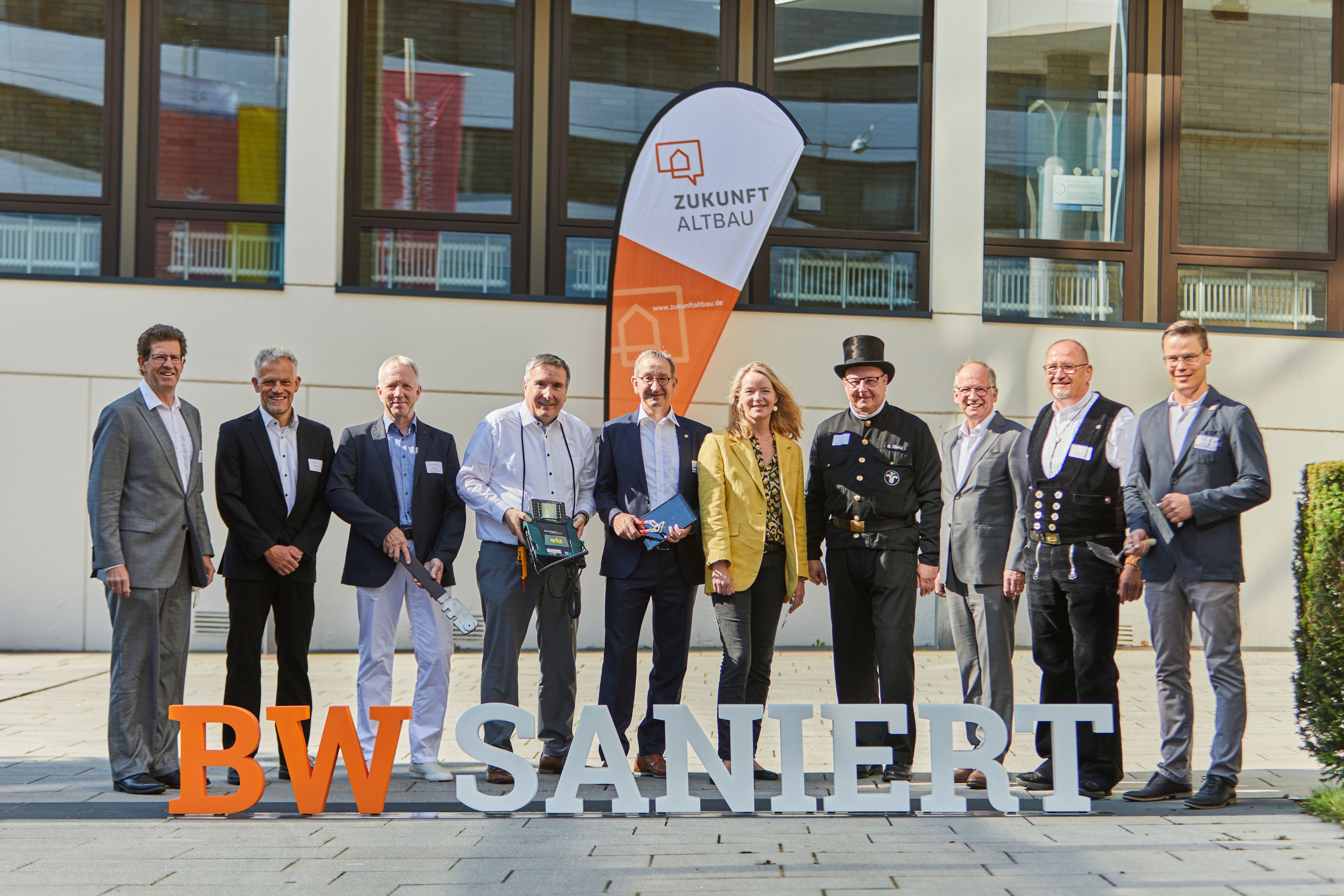Zehn Personen stehen vor einem Gebäude. Sie sind Teil der unterschiedlichen Klimagewerke. Vor den Personen stehen 3D-Buchstaben. Sie ergeben den Schriftzug "BW saniert"