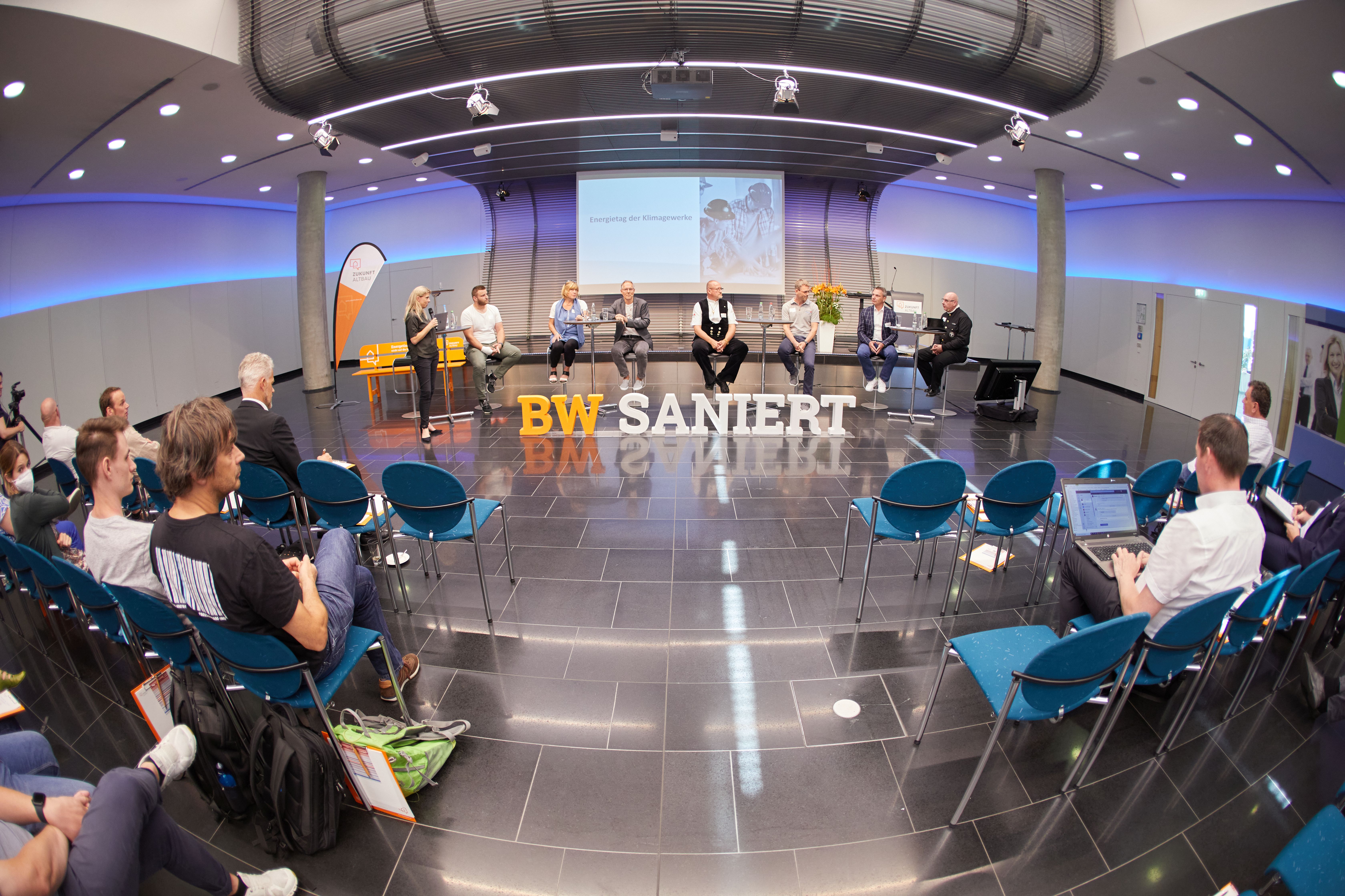 Teilnehmende beim Energietag für Klimagewerke sitzen in Richtung Bühne. Im Hintergrund ist eine Leinwand zu erkennen.