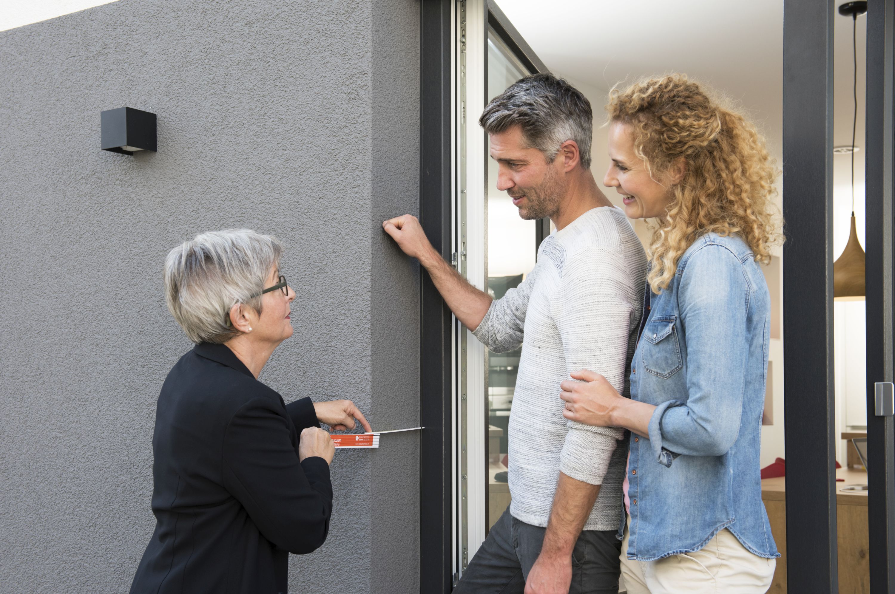 Bild zeigt ein junges Paar, das im Hauseingang steht, der Mann klopft an die Hauswand. Eine Energieberaterin steht links davon und zeigt mit einem Zollstock die Dicke der Fassadendämmung an.