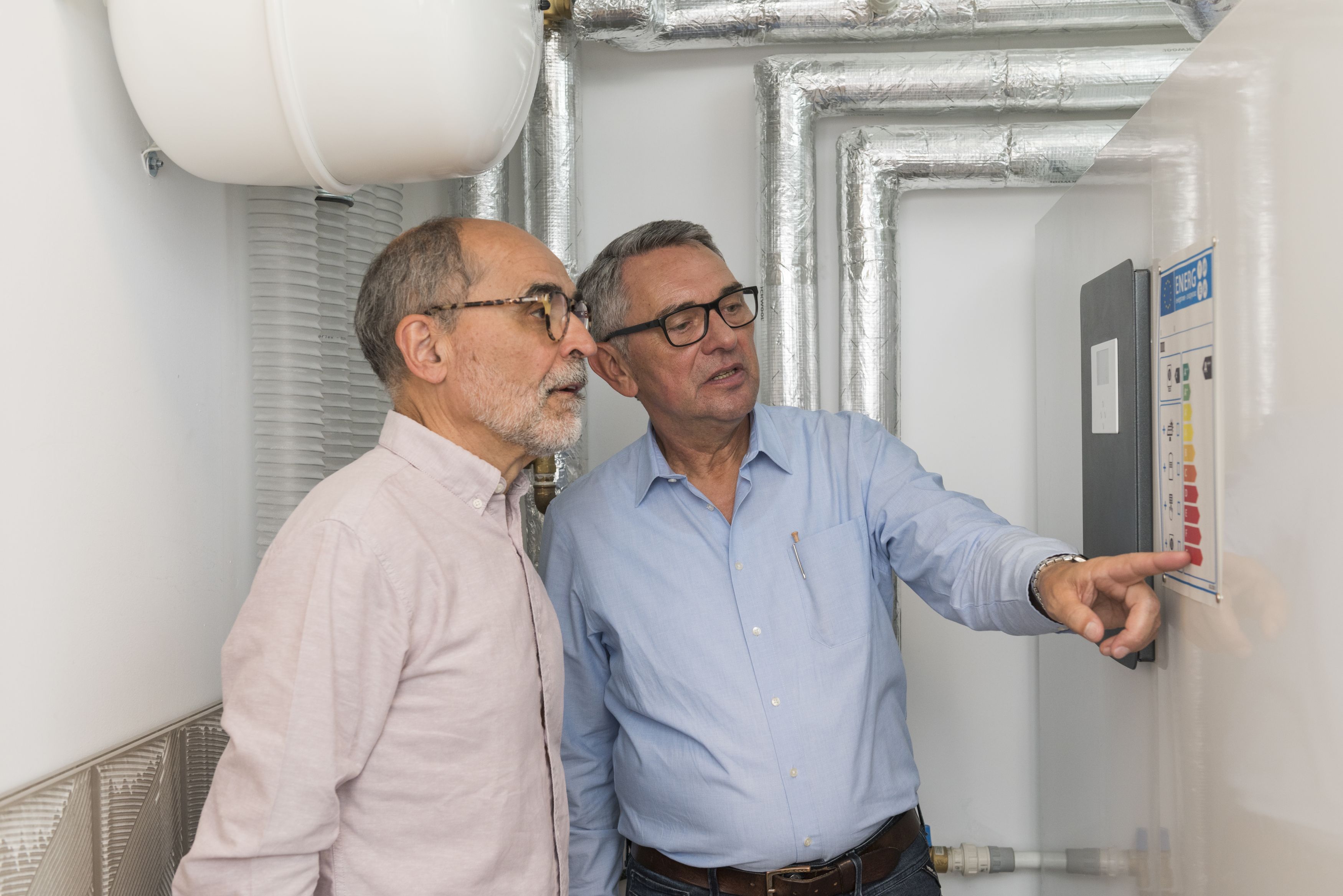 Bild zeigt zwei Männer im Heizungsraum. Der Energieberater rechts im Bild zeigt auf ein Erklärbild an der Wand, der Hauseigentümer schaut interessiert darauf.