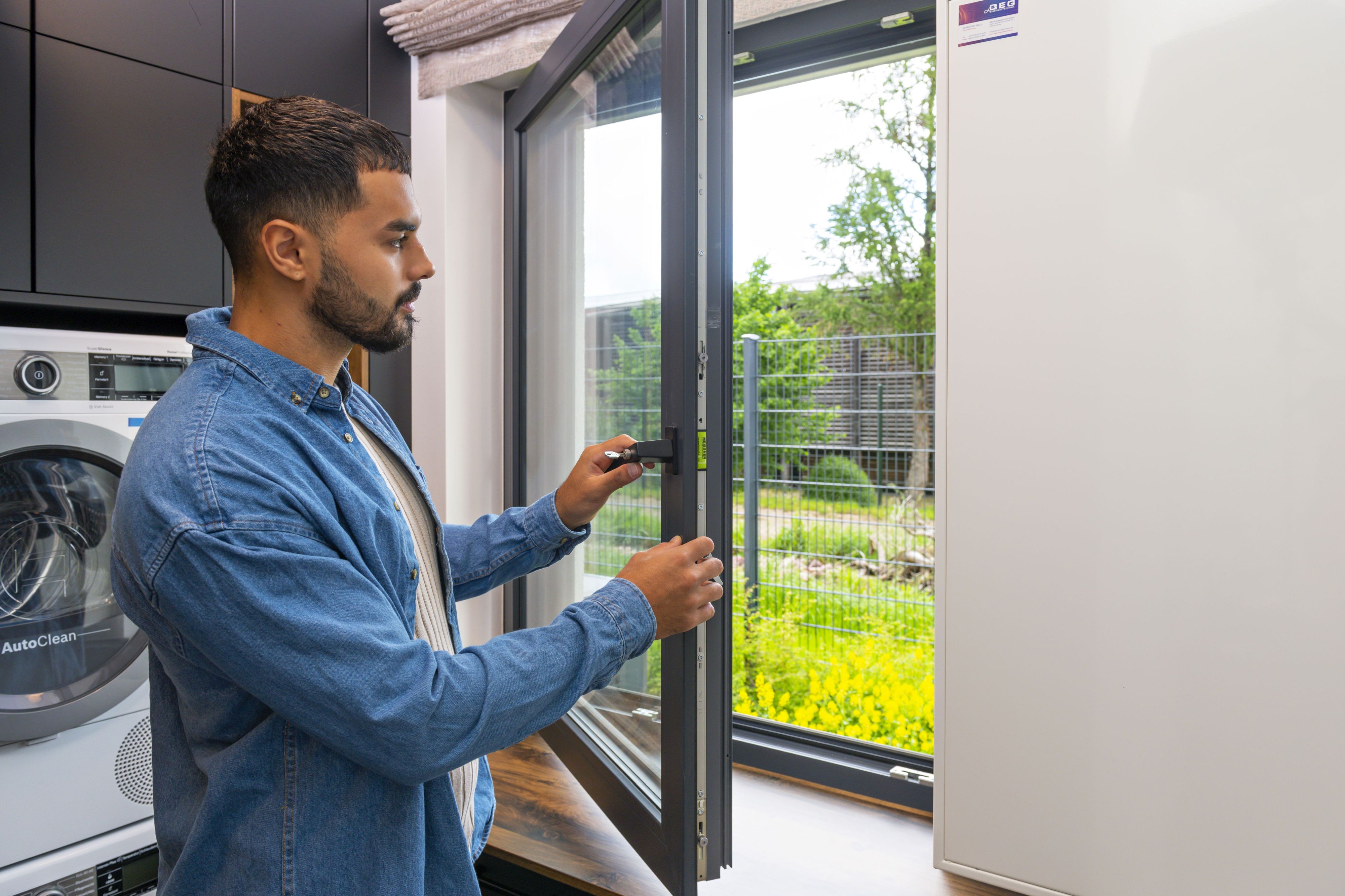 Auf dem Bild ist ein junger Mann in einem hellblauen Hemd zu sehen. Er steht vor einer Waschmaschine an einem Fenster, das er gerade öffnet. Draußen ist ein Stück eines Rasens zu erkennen
