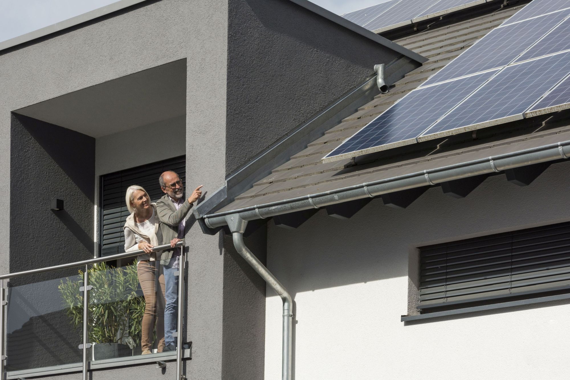 Bild zeigt ein Paar links im Bild, das auf einer Dachterasse steht. Sie schauen auf Photovoltaik-Module auf dem Dach, die sich rechts im Bild befinden.