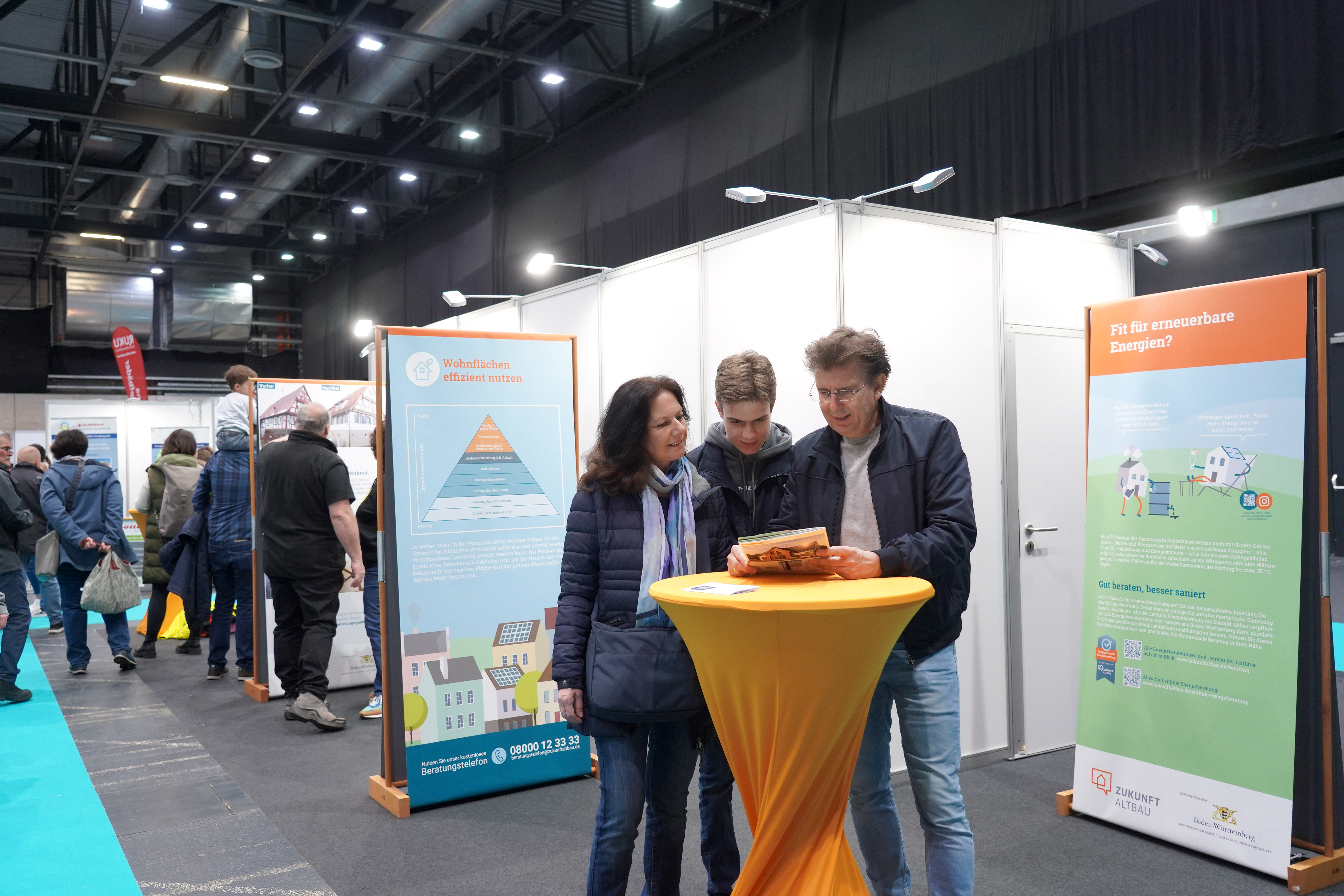 Das Foto zeigt eine Wanderausstellung. Drei Personen stehen um einen Stehtisch herum und unterhalten sich.