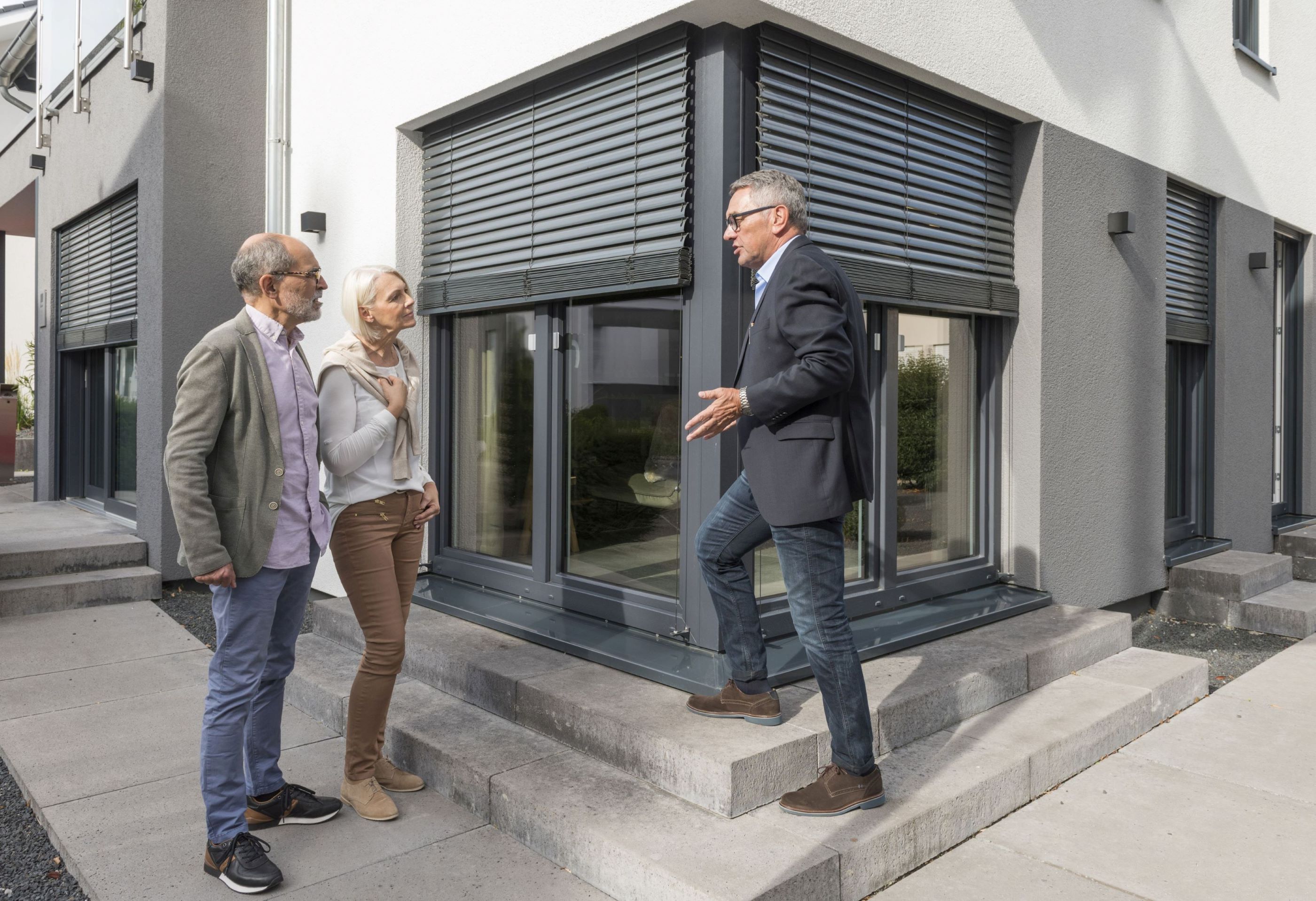 Das Bild zeigt drei Personen außerhalb eines Hauses direkt vor einer Balkontür und bodentiefen Fenstern. Der Gebäudeenergieberater erklärt einem Paar zeigt auf die heruntergefahrene Rolläden und erklärt, wie sie vor Hitze schützen.