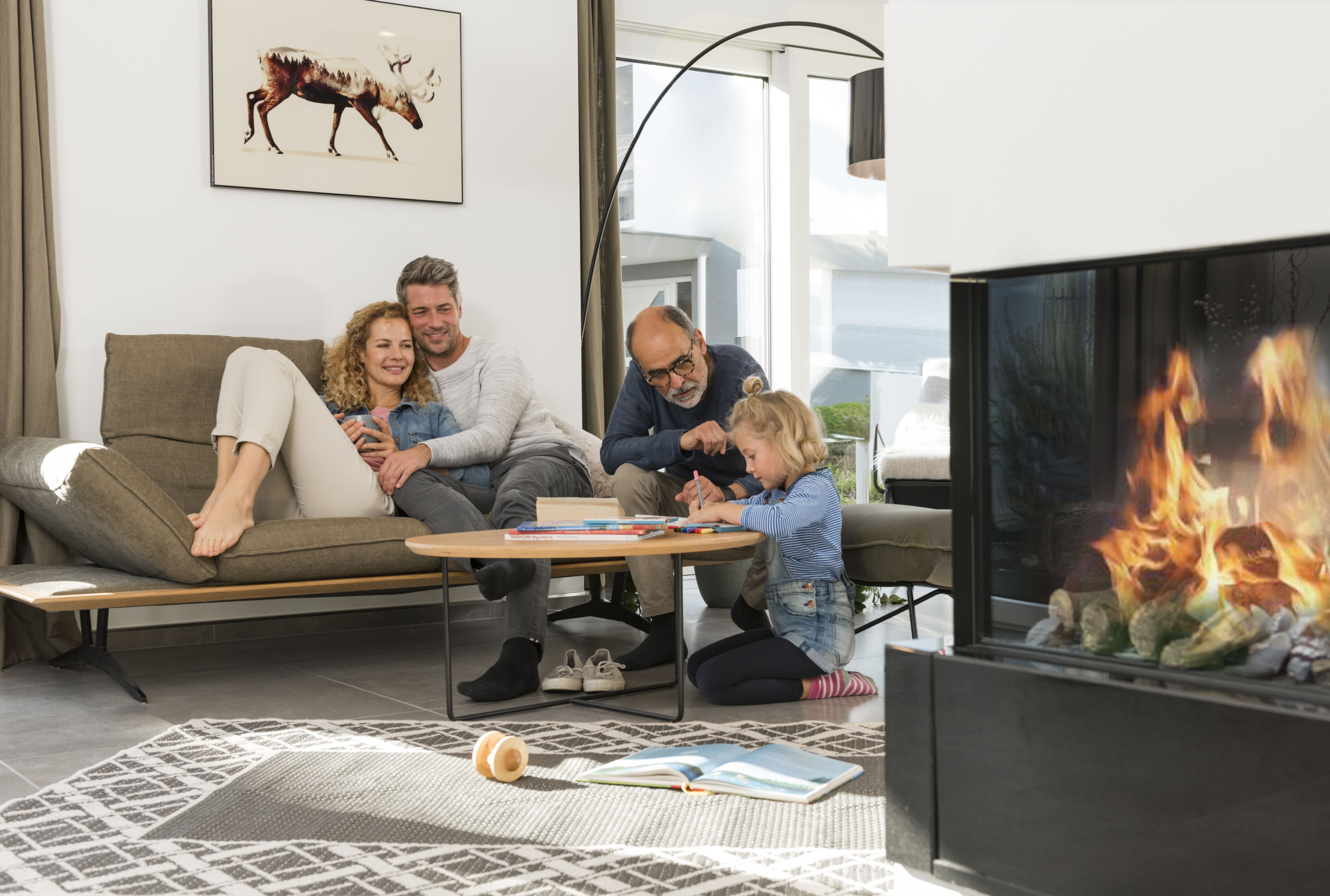 Bild zeigt eine junge Familie im Wohnzimmer. Das Paar sitzt gemeinsam auf dem Sofa, während der Opa mit dem Enkel spielt.